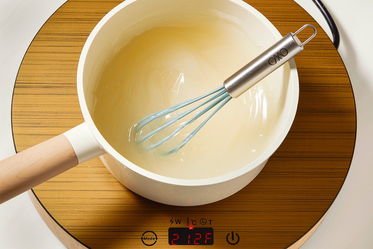 White saucepan with a light wooden handle on an induction cooktop, containing a lemon curd being stirred at 212°F with a whisk.