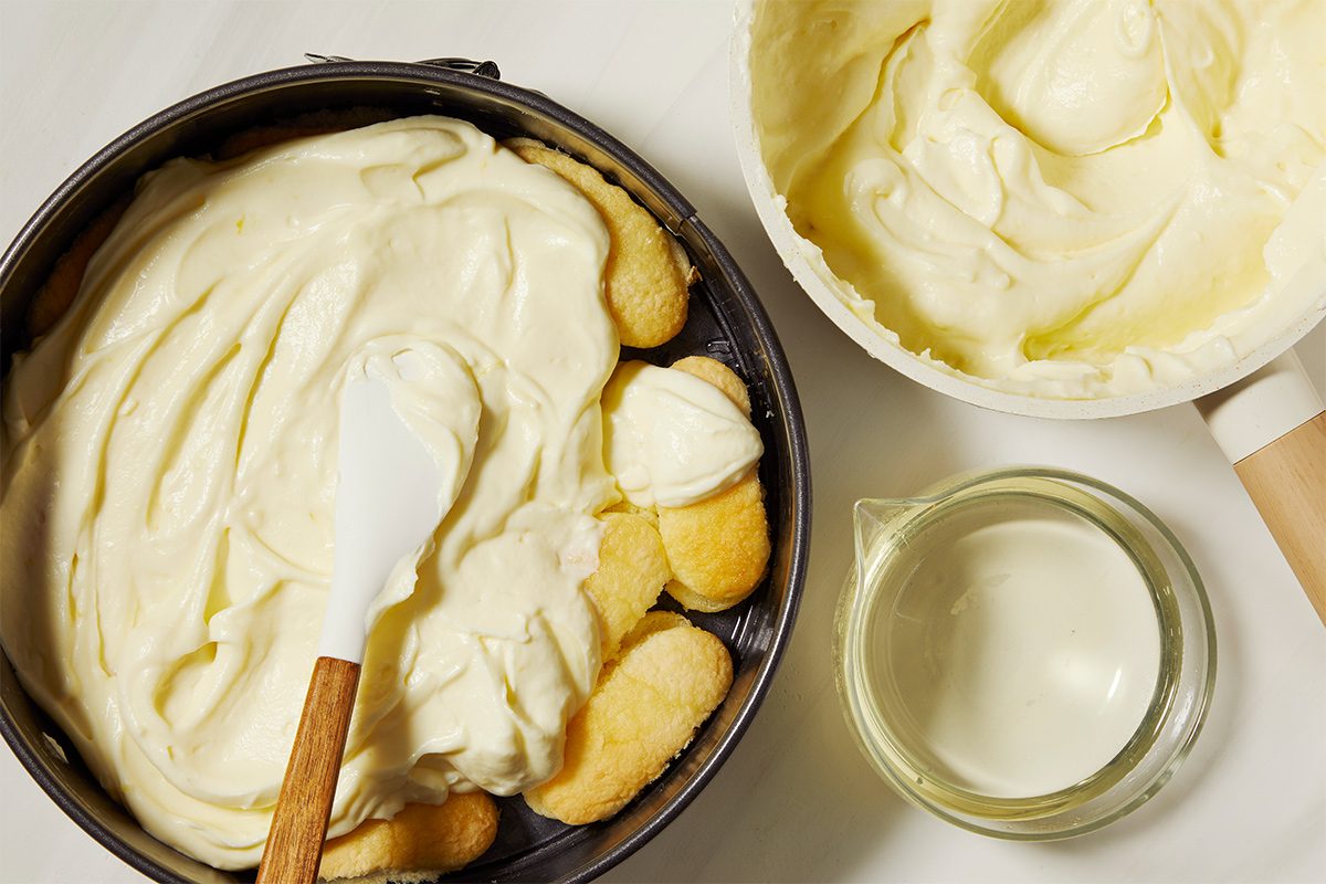 A baking tin is filled with ladyfingers and creamy mixture, being spread with a spatula. Nearby is a bowl of the same cream and a small jug of limoncello.