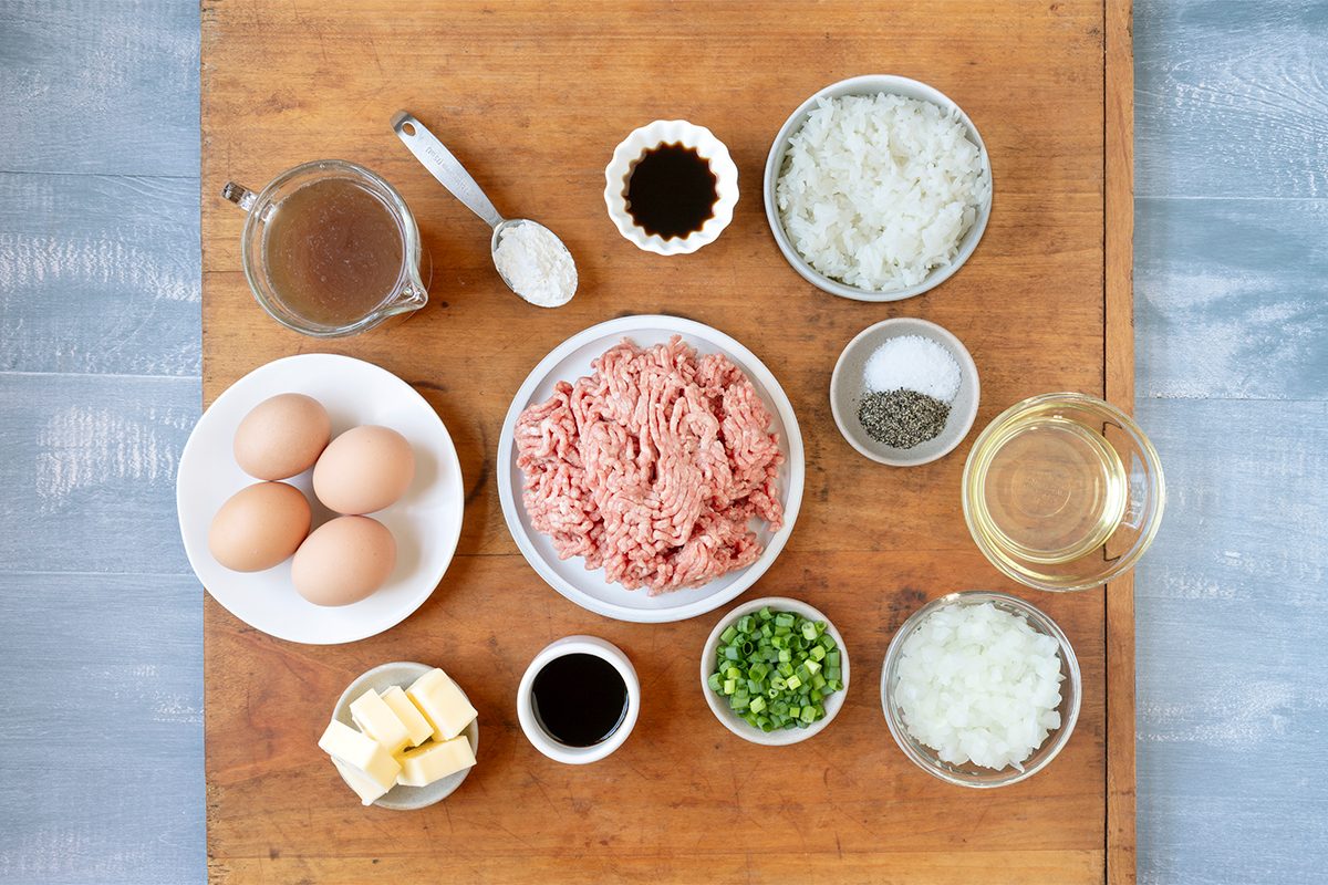 Ingredients neatly arranged on a wooden surface, including ground meat, eggs, chopped green peppers, diced onions, butter, sauces, seasoning, and liquids in small bowls and plates.