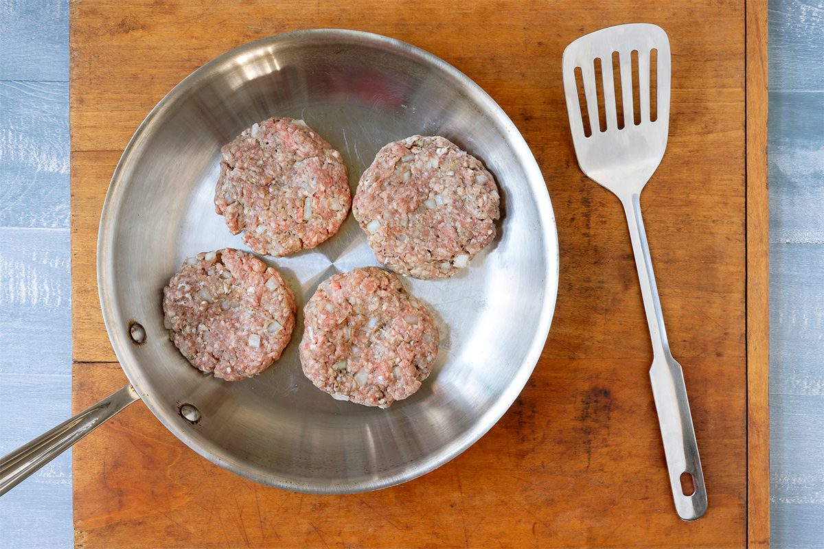 Four raw burger patties are placed in a stainless steel frying pan on a wooden cutting board. A metal spatula lies next to the pan. The background is a light blue surface.