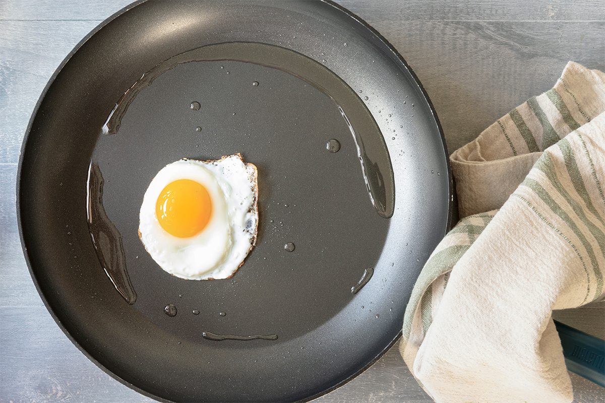 A single sunny-side-up egg is frying in a black non-stick pan on a gray surface. A striped cloth is placed nearby.