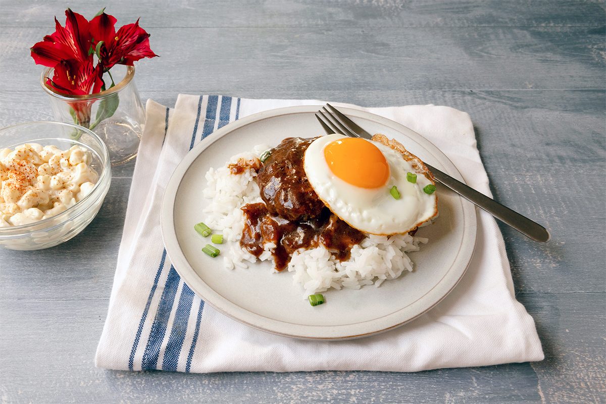 A plate of rice topped with a fried egg and brown sauce, garnished with green onions, set on a striped napkin. A fork rests on the plate. A bowl of crumbled topping and a red flower in a glass are beside it on a wooden surface.