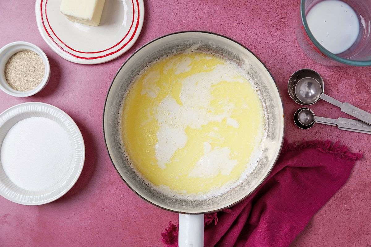 A mixing bowl with a creamy yellow batter on a pink surface, surrounded by ingredients like butter on a plate, a bowl of sugar, measuring cups, and a red cloth.
