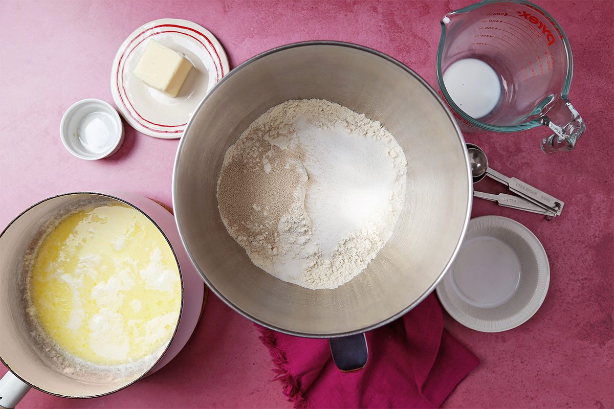 A pink surface displays bowls and ingredients for baking: a large bowl with flour and sugar, a small pitcher with liquid, a partial bowl with melted butter, and small dishes with butter and other ingredients. A red cloth is partially visible.