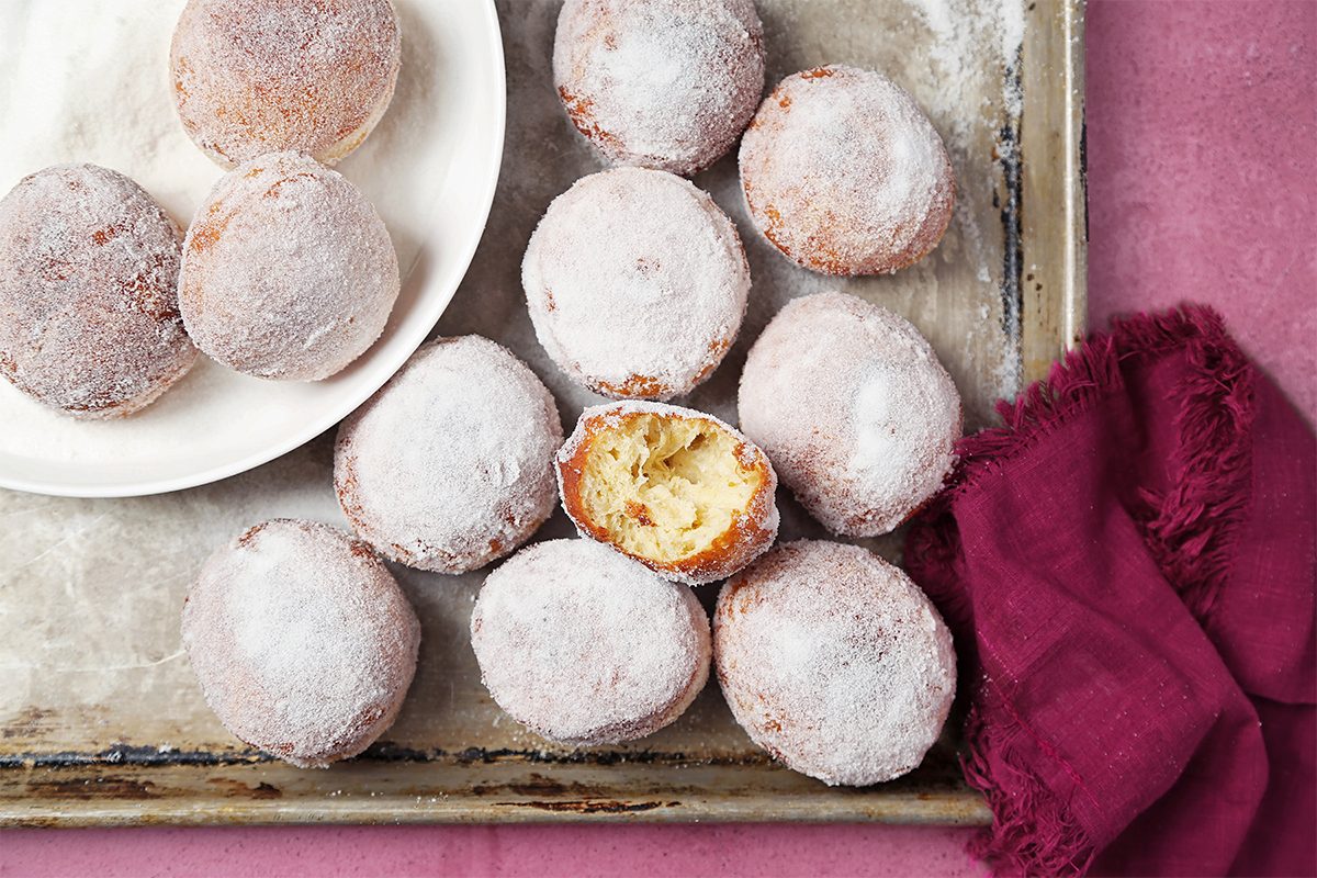 Powdered sugar-covered doughnuts arranged on a tray, with one cut open to reveal the inside. A few doughnuts are placed on a white plate on the tray. A red cloth is draped on the right side. The background is pink.