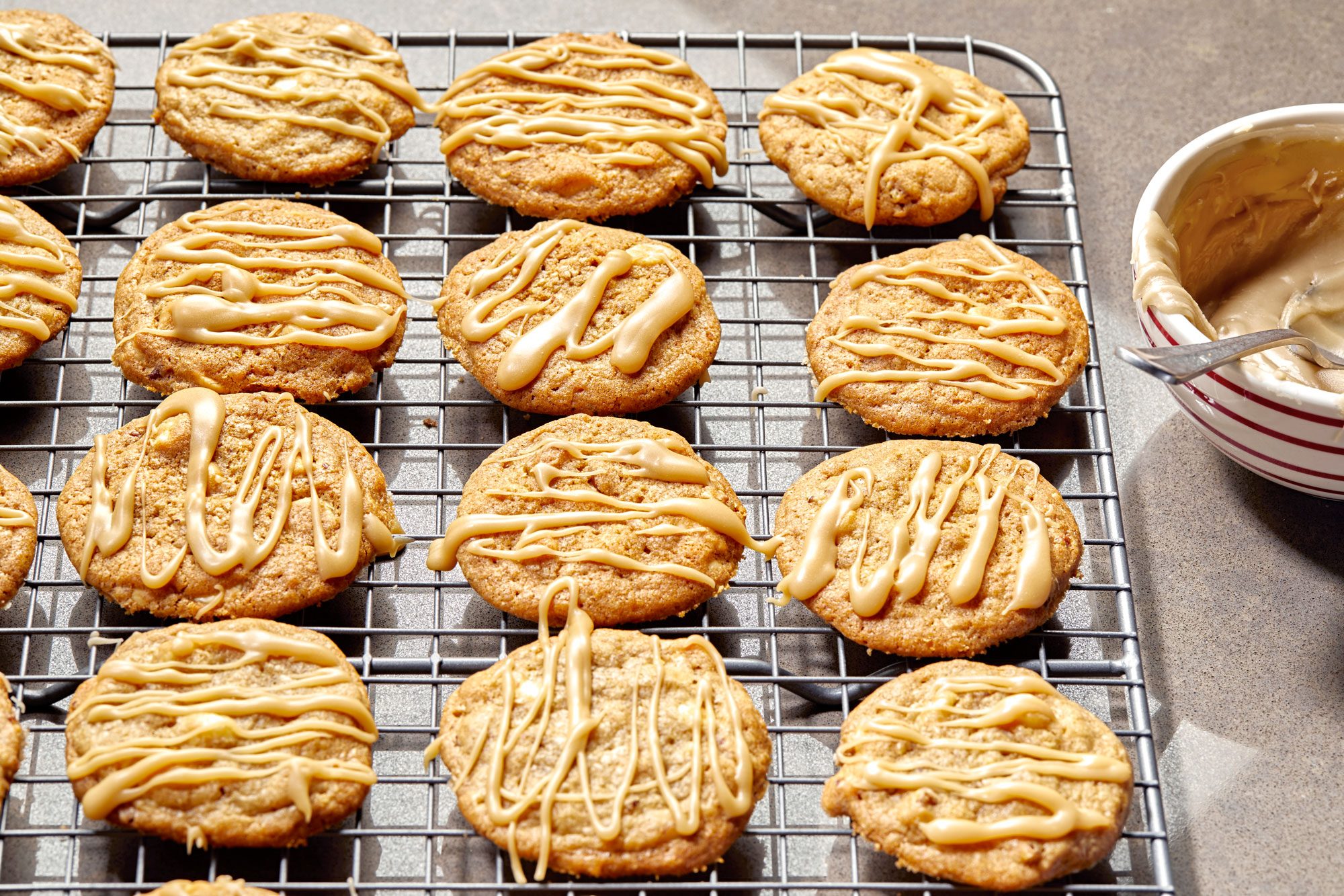 Horizontal AP Pull shot of a cooling rack with freshly baked cookies; the cookies are iced with a caramel glaze, and the glaze is still wet and shiny; a small bowl of caramel glaze is on the right side of the image, and a spoon is in the bowl