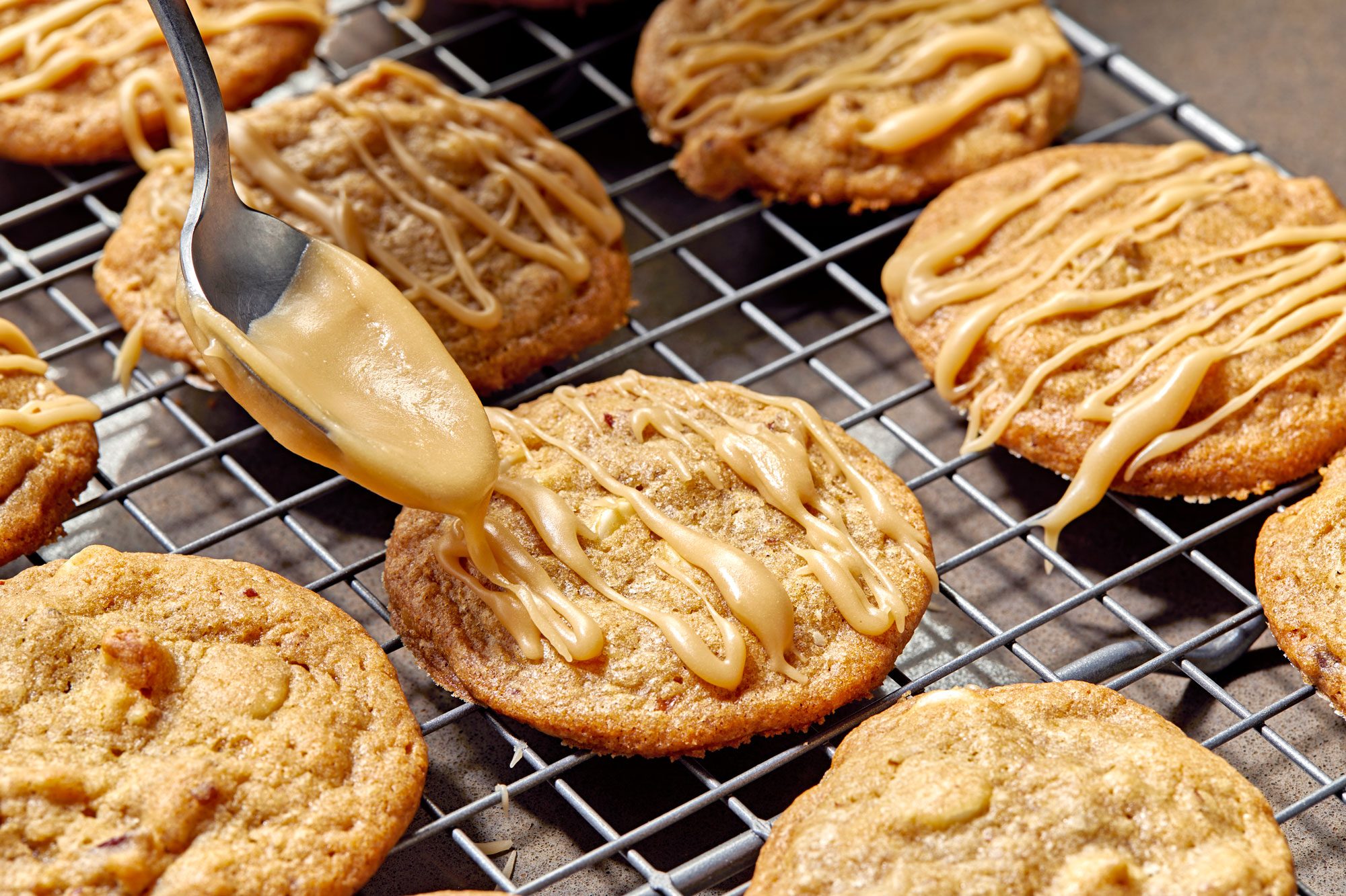 a close-up of a baking rack with golden-brown cookies being drizzled with caramel icing; a spoon is pouring the icing over one of the cookies