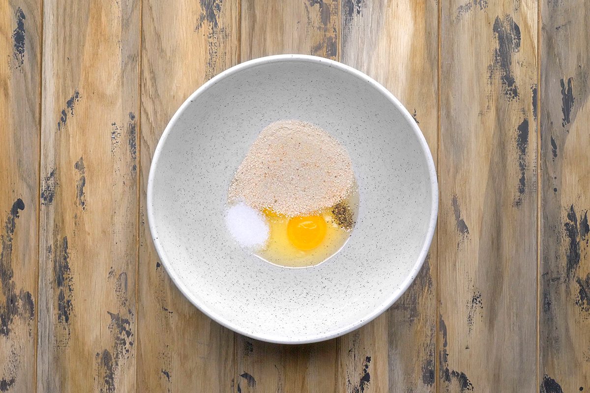 A white bowl on a wooden surface contains a raw egg, a mound of flour, a small pile of sugar, and a dash of salt, ready to be mixed.