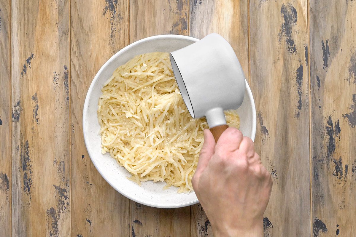 Person pouring shredded potatoes from a cup into a white bowl on a wooden surface.