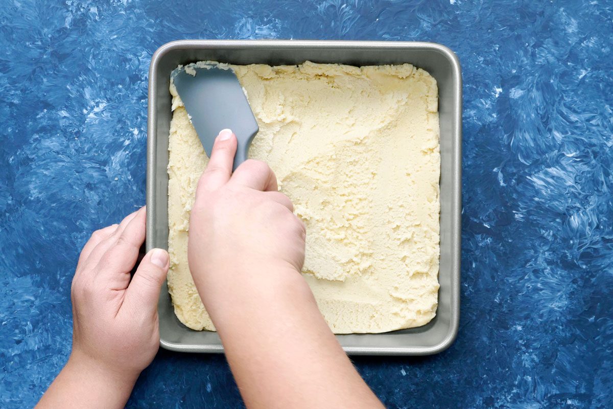 overhead shot of a person spreading a white, dough-like substance in a rectangular baking pan with a blue spatula; the pan is on a blue textured surface