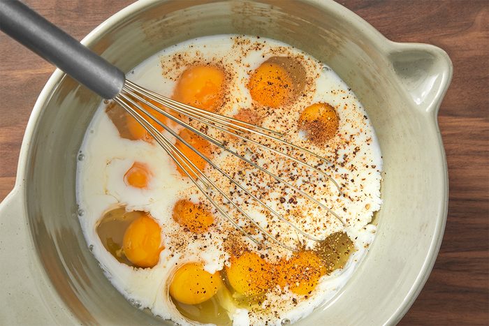 A bowl containing several raw eggs, milk, and a sprinkle of pepper, with a whisk partially submerged in the mixture, resting on a wooden surface.