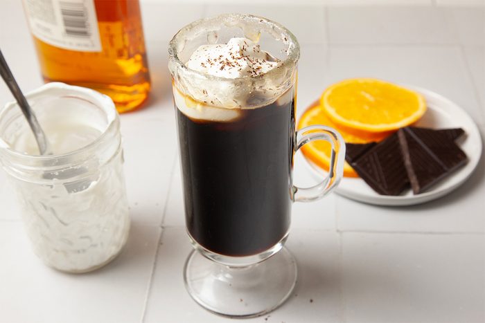 A glass of Irish coffee topped with whipped cream and chocolate shavings on a table. Nearby, there's a jar with a spoon, a bottle, and a plate with orange slices and dark chocolate squares.