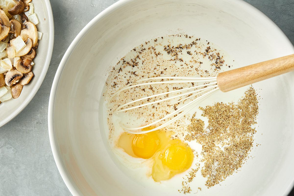 A mixing bowl contains two cracked eggs, ground spices or herbs, and a whisk. Partially visible on the side is a plate with sliced mushrooms and onions. The bowl is set on a gray surface.