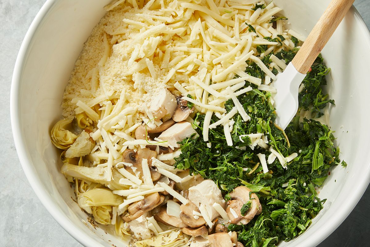 A bowl contains shredded cheese, sautéed spinach, sliced mushrooms, artichoke hearts, and grated parmesan. A spatula is partially mixing the ingredients, ready for preparation.