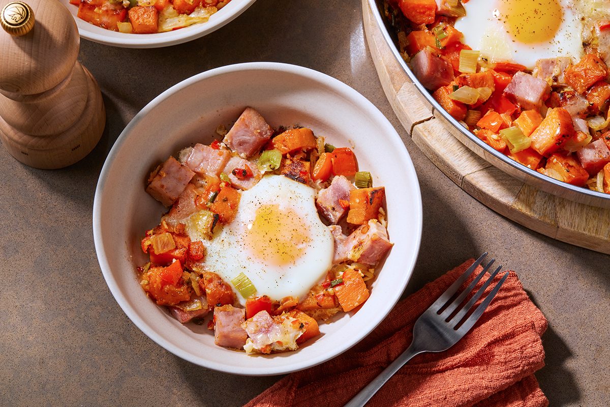 A bowl of hash with diced ham, sweet potatoes, and vegetables, topped with a fried egg. It's served on a brown tabletop alongside a fork on an orange napkin and a wooden pepper mill. Another dish is partially visible in the background.