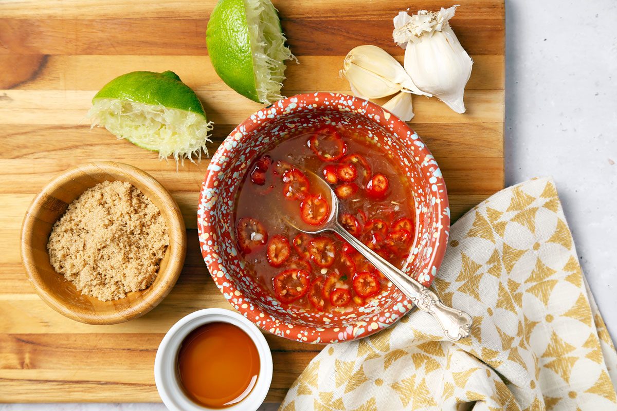 A vibrant, textured sauce with red chili slices in a red-and-white bowl on a wooden cutting board. Surrounding items include garlic cloves, squeezed lime halves, a small bowl of brown sugar, and a cup of soy sauce, alongside a patterned napkin.