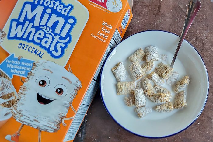 A bowl of Frosted Mini-Wheats with milk and a spoon sits beside an open box of the cereal on a countertop. The cereal box displays a smiling wheat character.