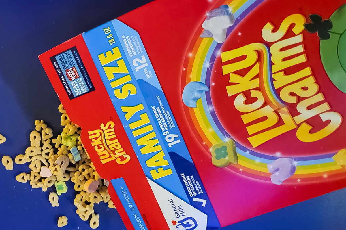 A box of Lucky Charms cereal labeled "Family Size" is tilted on a surface, spilling cereal pieces. The box features colorful packaging with the cereal