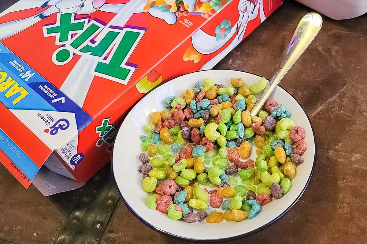 A bowl of colorful Trix cereal with milk and a silver spoon next to a Trix cereal box on a wooden table.