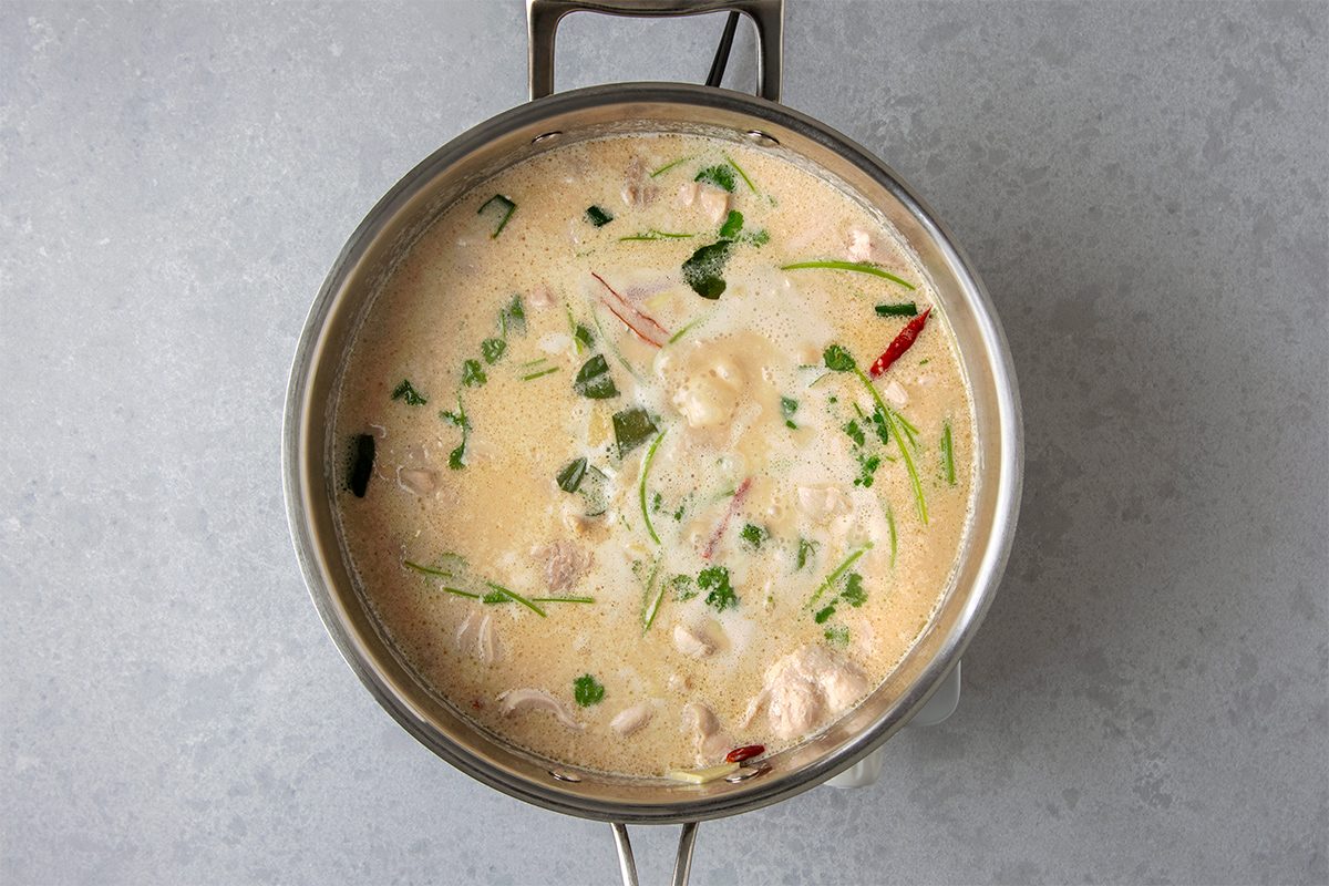 A pot of creamy soup with herbs, red chili slices, and pieces of chicken or seafood, possibly a Thai coconut soup, on a gray background.