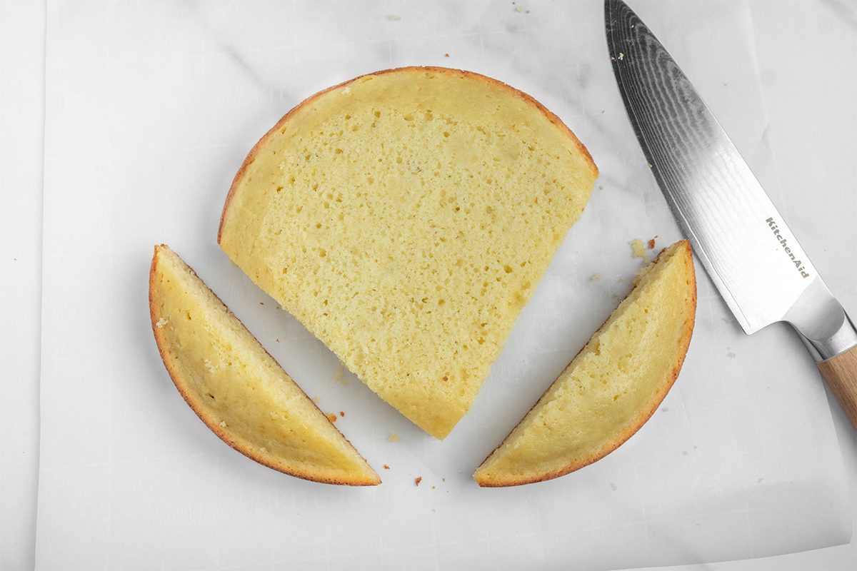 A round cake is cut into three pieces on a white surface. Two smaller, wedge-shaped pieces flank a larger semicircular piece. A silver knife with a wooden handle is placed beside them.