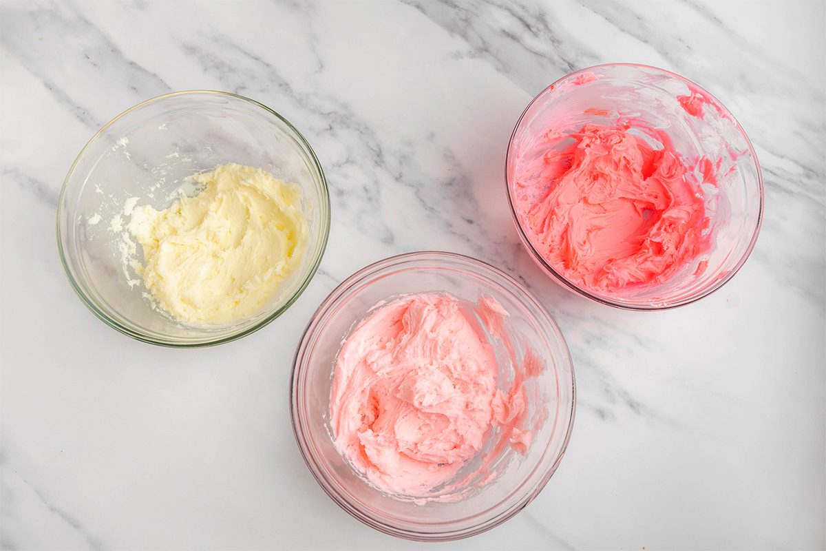 Three bowls containing whipped cream on a marble surface. The bowls have creams in pastel shades of white, light pink, and bright pink.
