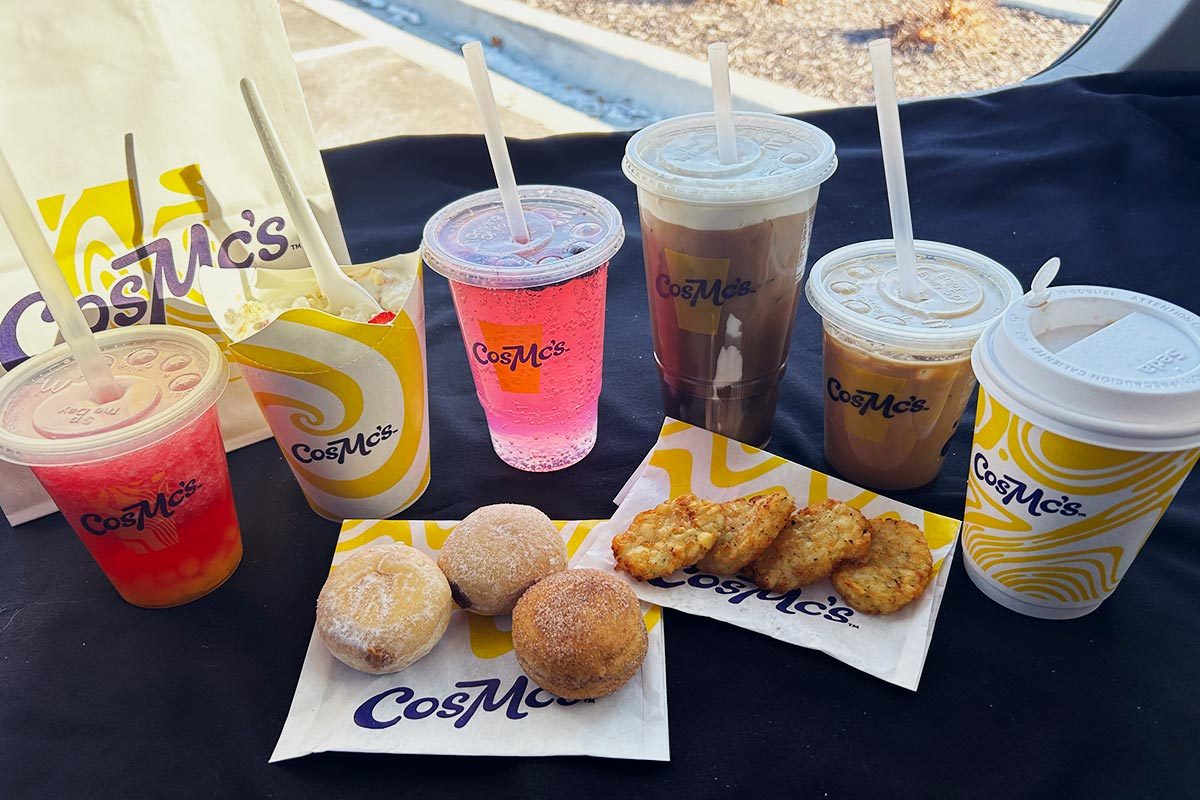 Six CosMc's drinks, including iced beverages and hot coffees, filled mini donuts, and hash browns on a black surface with a CosMc's bag in the background