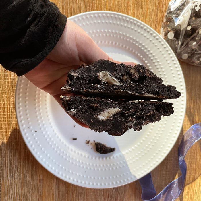Hand holds a cross section of a dark chocolate cookie with toffee chips above a white plate and wooden table