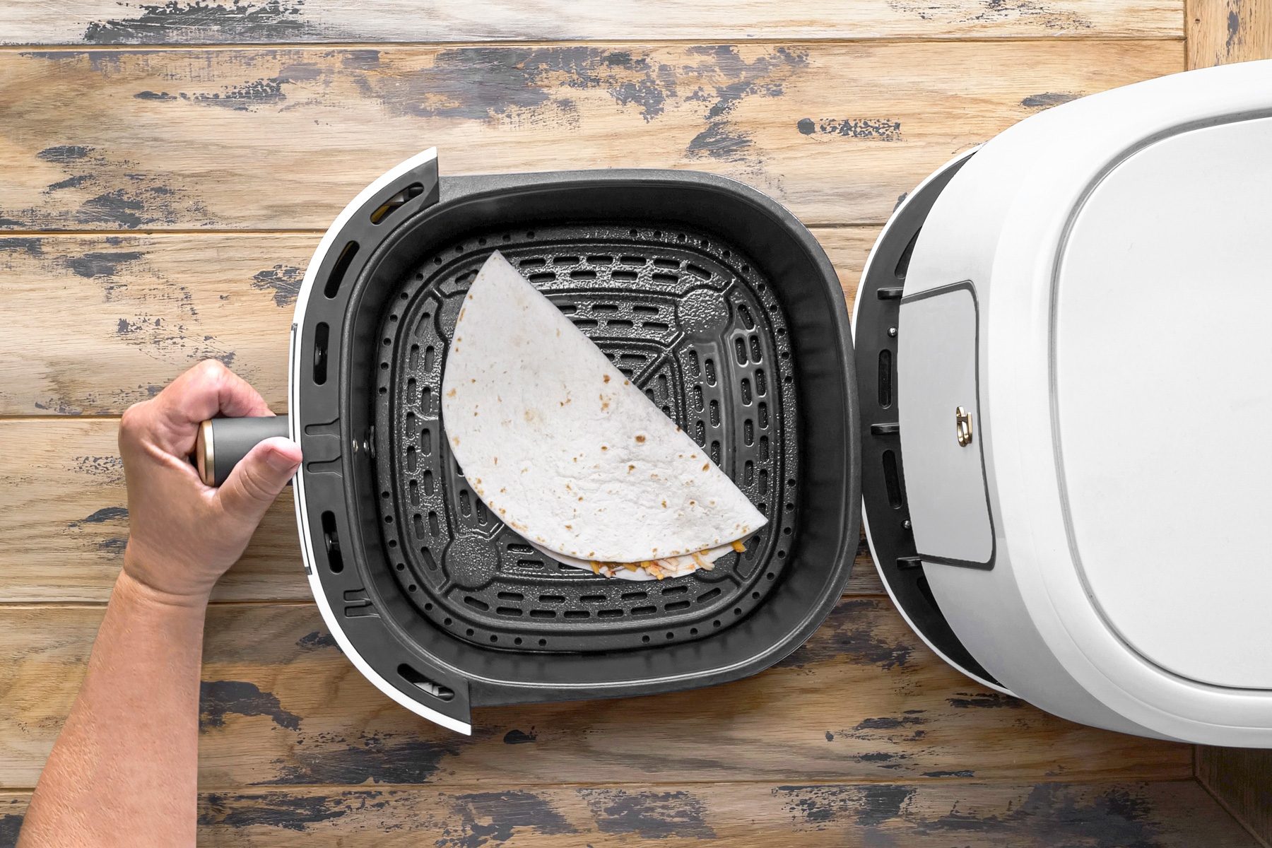 overhead shot of a hand is holding the handle of an air fryer basket; the basket is open; inside the basket is a folded white tortilla; the tortilla is filled with cheese and other ingredients; the basket is sitting on a wooden table