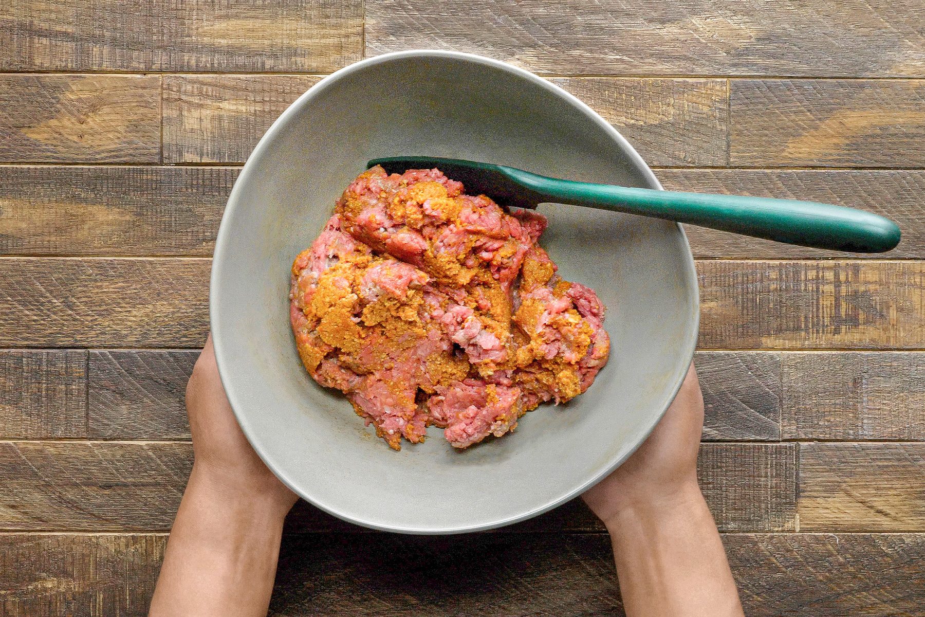 overhead shot of a person holding a grey bowl containing raw ground meat; a green spatula is also in the bowl; the bowl is sitting on a wooden surface