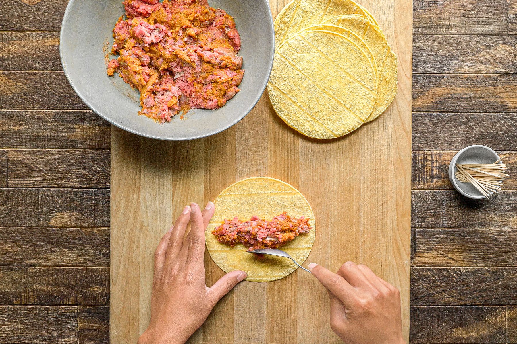 overhead shot of a person preparing to make a taco; the person has a cutting board in front of them with a stack of tortillas on the left side of the board and a single tortilla with ground meat on the right side of the board; the person is using a spoon to spread the ground meat on the tortilla; a gray bowl with ground meat is on the left side of the image