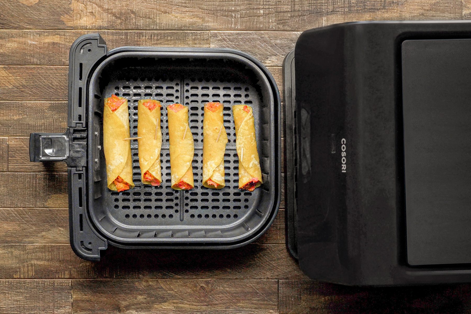 overhead shot of an air fryer with the basket pulled out, revealing five rolled up tortillas, the air fryer is sitting on a wooden table.