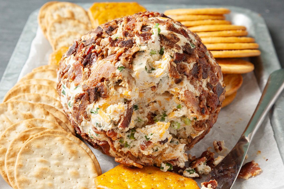 Close shot of Bacon Ranch Cheese Ball; on a platter with assorted crackers; knife; grey surface