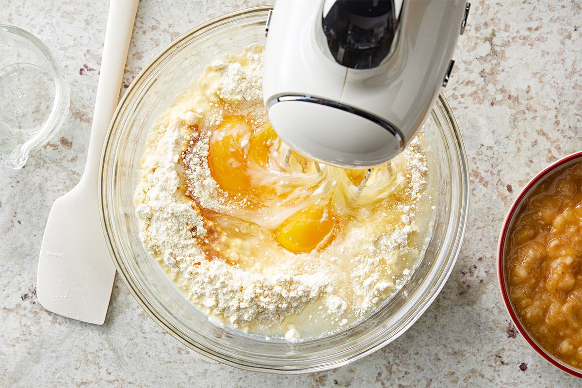 overhead shot of a mixing bowl filled with a batter that includes eggs and flour being blended with a hand mixer; the mixer is partially submerged in the mixture, beside the bowl, there is a small container a mixture; the countertop is a light colored surface