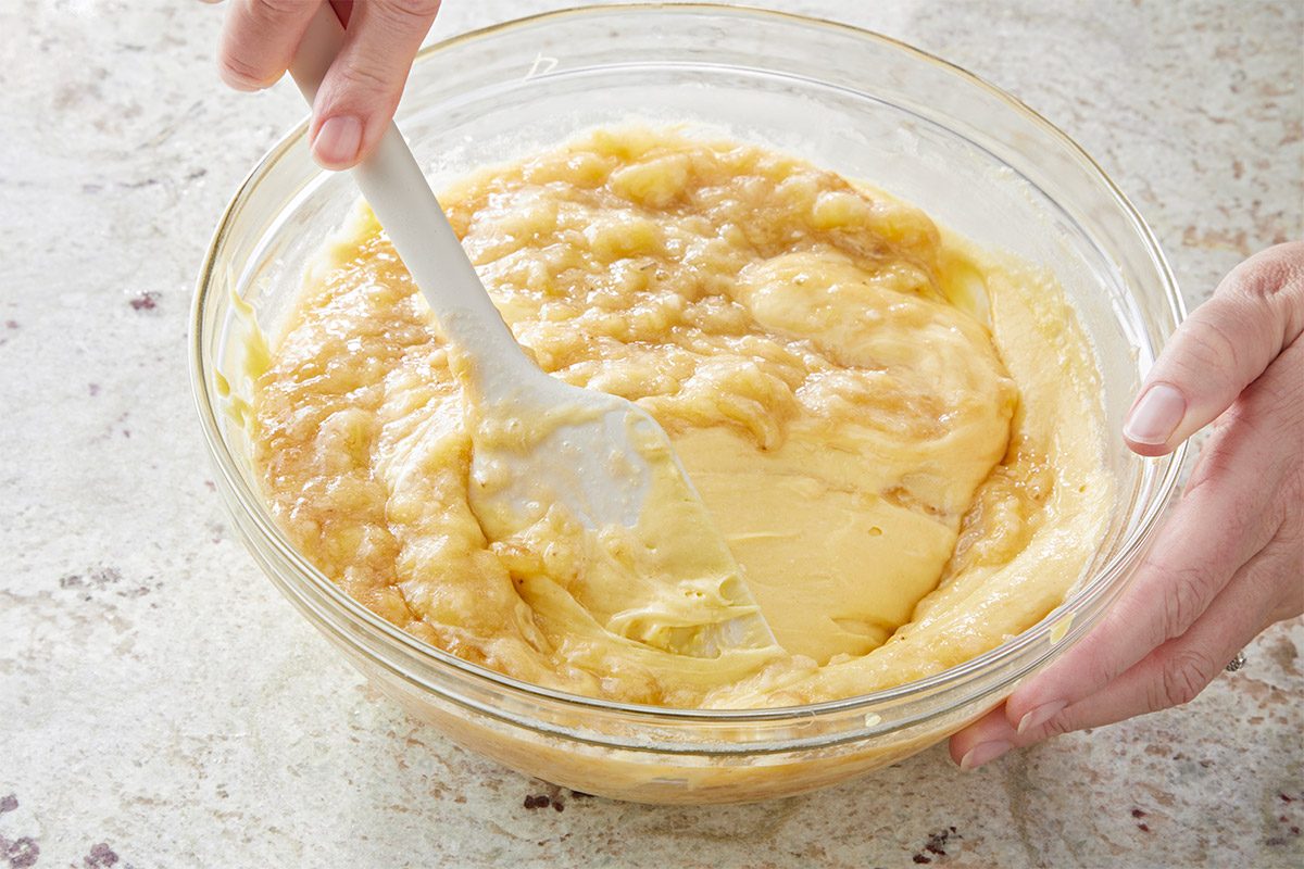 3/4th shot of a clear, round bowl containing a creamy, yellowish a batter, one hand is holding the bowl while another hand is using a spatula to stir the mixture; the background is lightly textured surface
