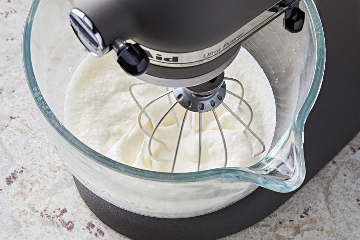 3/4th shot of a stand mixer with a glass mixing bowl, showcasing the mixture inside, which is a whipped cream, the stand mixer is positioned at an angle, with its stainless steel mixing attachment visible, the surface beneath the bowl has a marble like texture