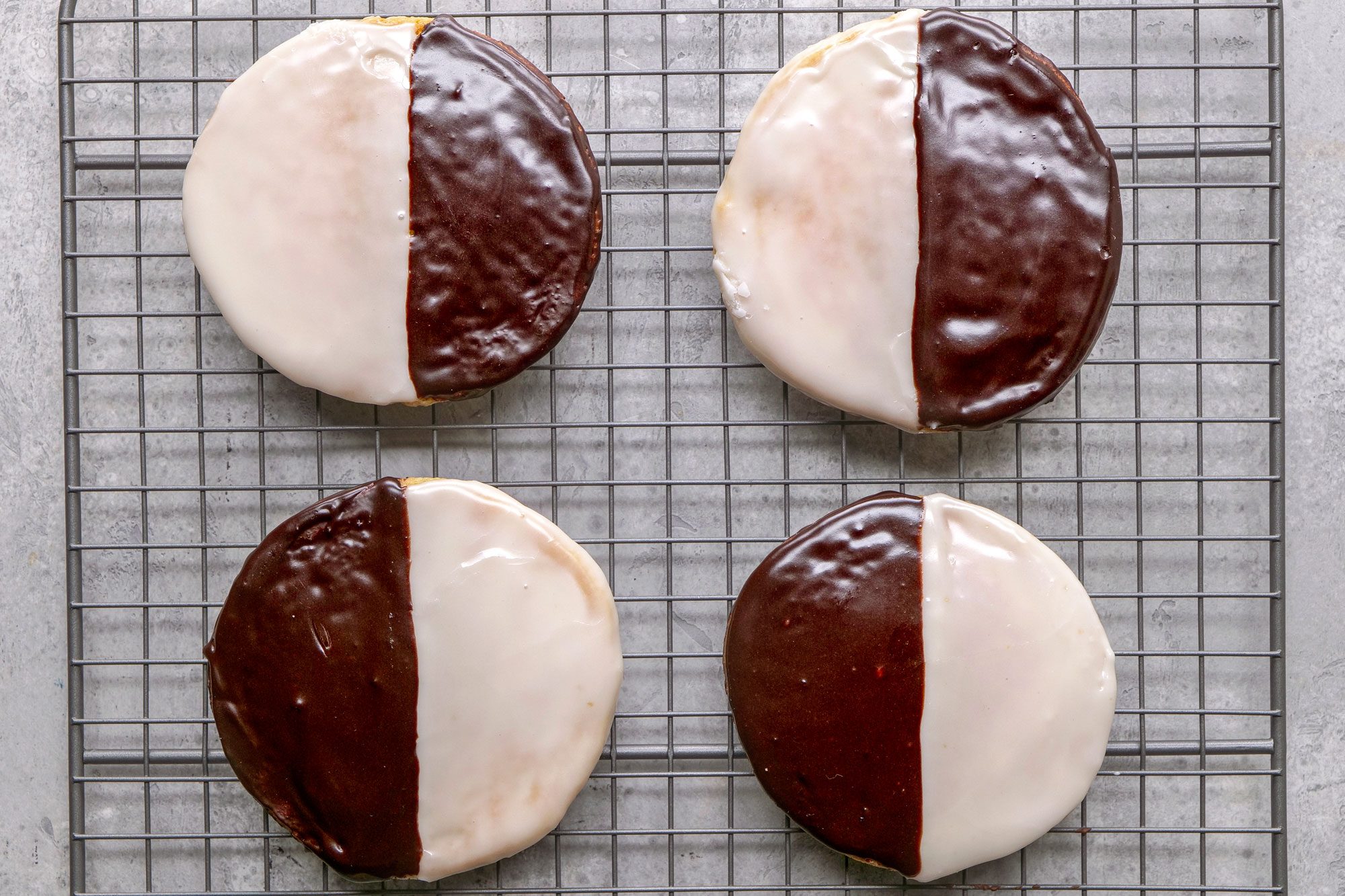 overhead shot of Four black and white cookies are arranged on a grey wire cooling rack, the background is a plain grey surface