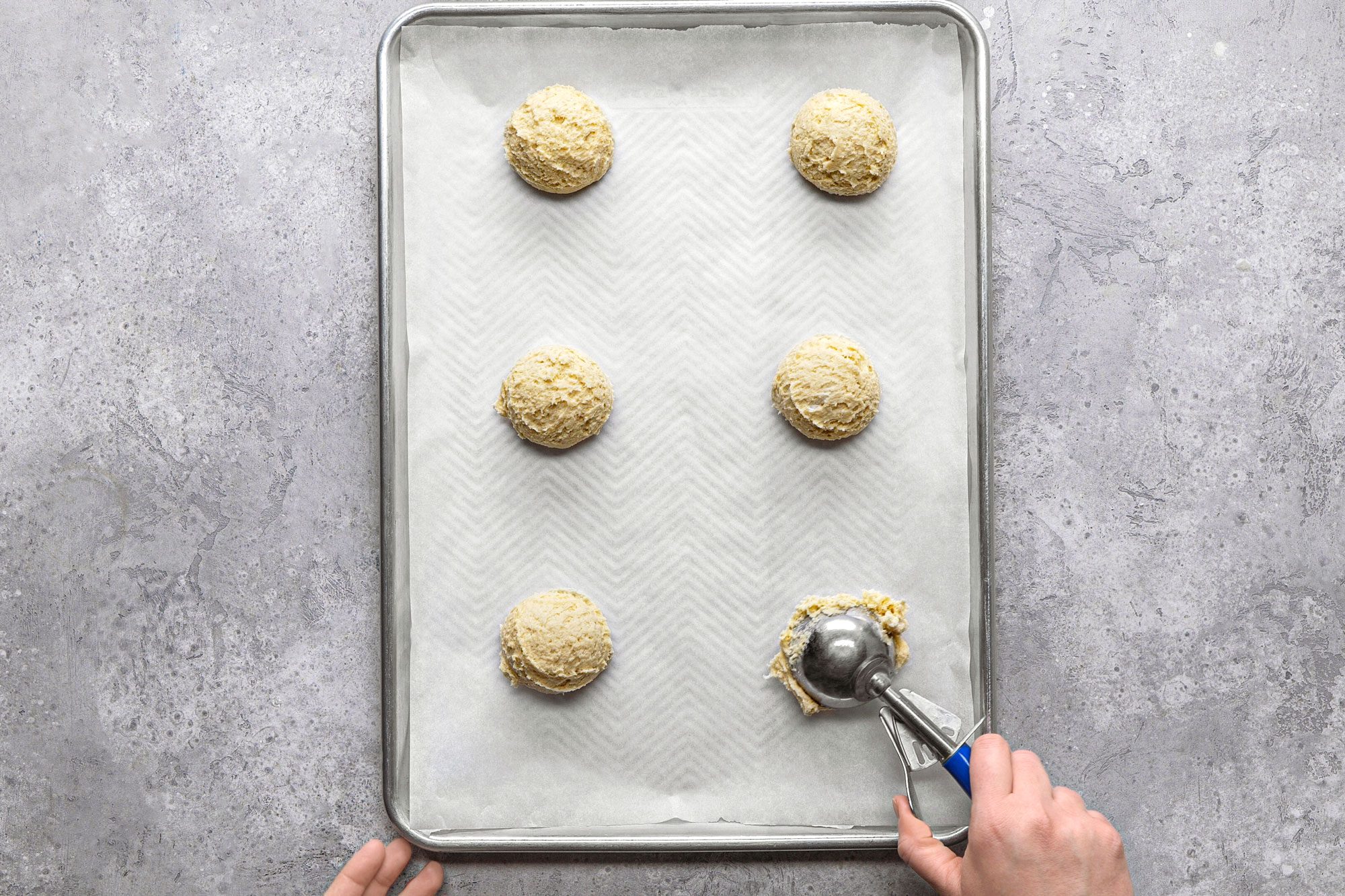 overhead shot of a baking sheet lined with white parchment paper, on which five mounds cookie dough are neatly arranged, a silver cookie scoop is in the process of placing another scoop of dough onto the sheet, and the background is a grey