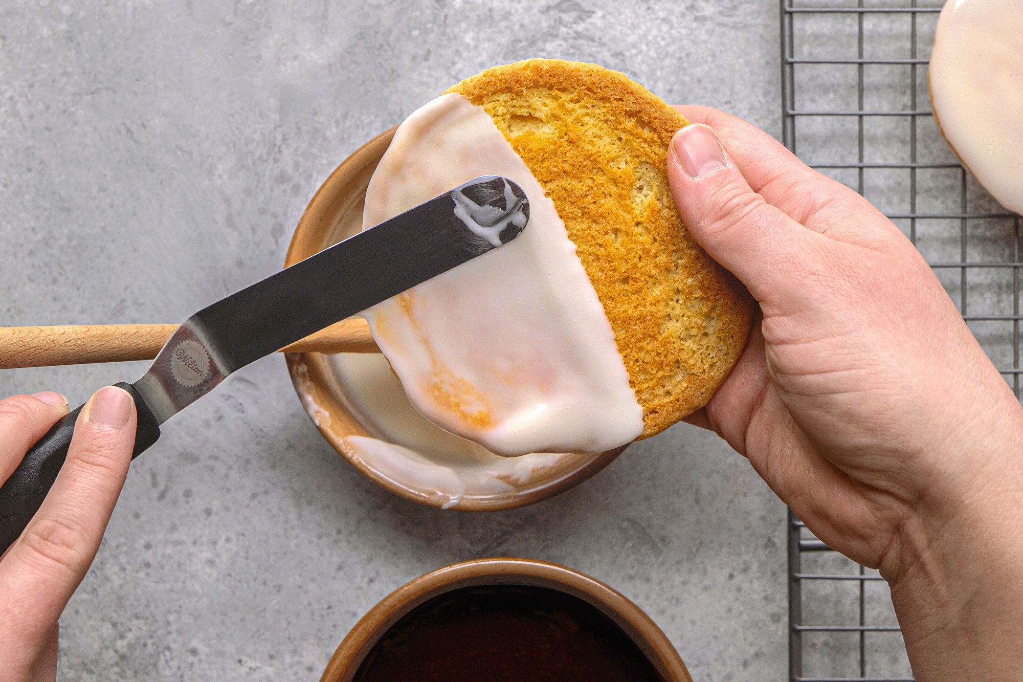 overhead shot of hands using a spatula to apply white frosting to a round cookie, a bowl containing more white frosting is visible below the cookie, and a bowl of dark, chocolate frosting is positioned at the bottom of the frame, the background is a textured grey surface