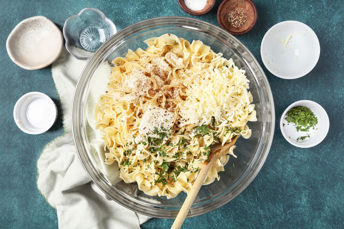 Overhead shot of cooked noodles in a large glass bowl; immediately add the remaining ingredients and toss to coat; spatula; napkin; teal green surface;