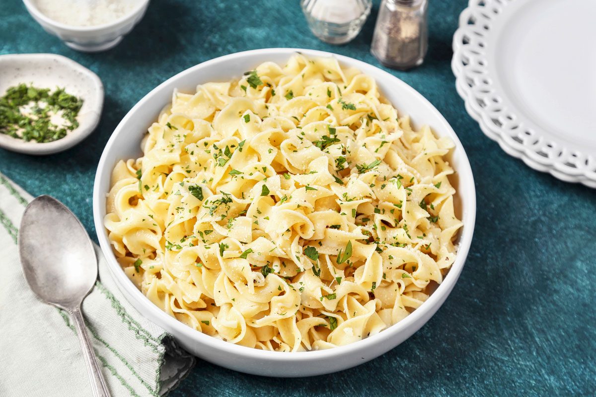 Table view shot of Buttered Noodles; In a large white bowl; serving plates; serving spoon; napkin; teal green surface;
