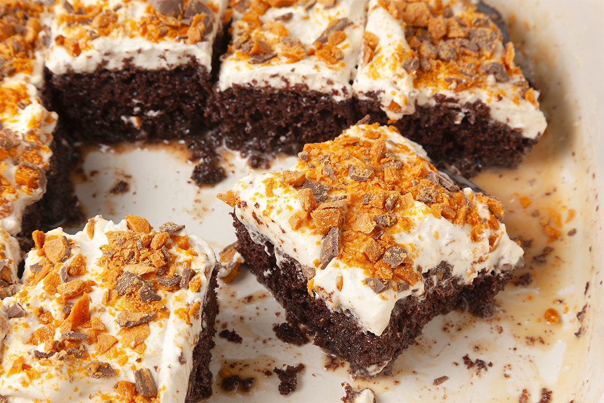 Close-up of a sliced chocolate cake with white frosting and crumbled toffee bits on top. The cake is partially cut, revealing moist, rich layers.