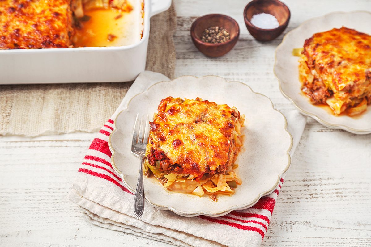A serving of lasagna on a scalloped white plate with a fork beside it. The plate is set on a red and white striped cloth. A dish in the background contains the remaining lasagna. Two small bowls hold salt and pepper.