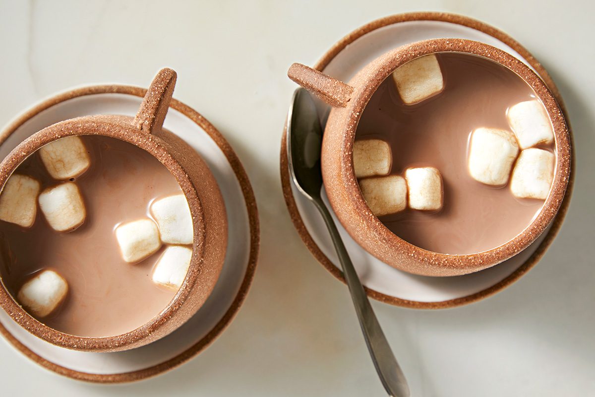 Two cups of hot chocolate topped with marshmallows sit on saucers. A spoon rests on one saucer. The cups and saucers are a matching light brown color, and the scene is set on a light-colored surface.
