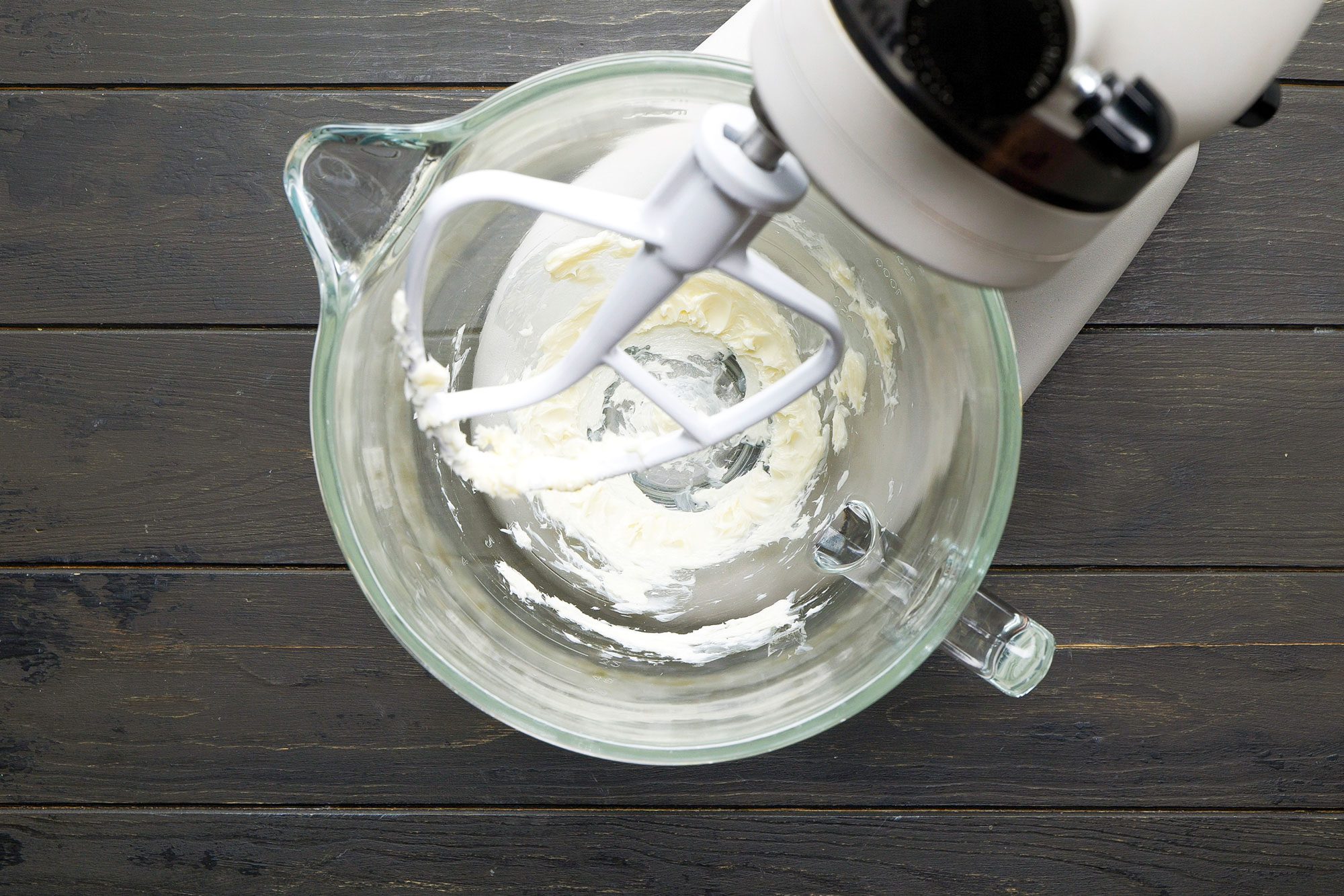 overhead shot of a clear mixing bowl that appears to have remnants of a creamy mixture stuck to its sides; a white mixing attachment is visible, showing the process of mixing ingredients; the bowl sits on a dark wooden surface