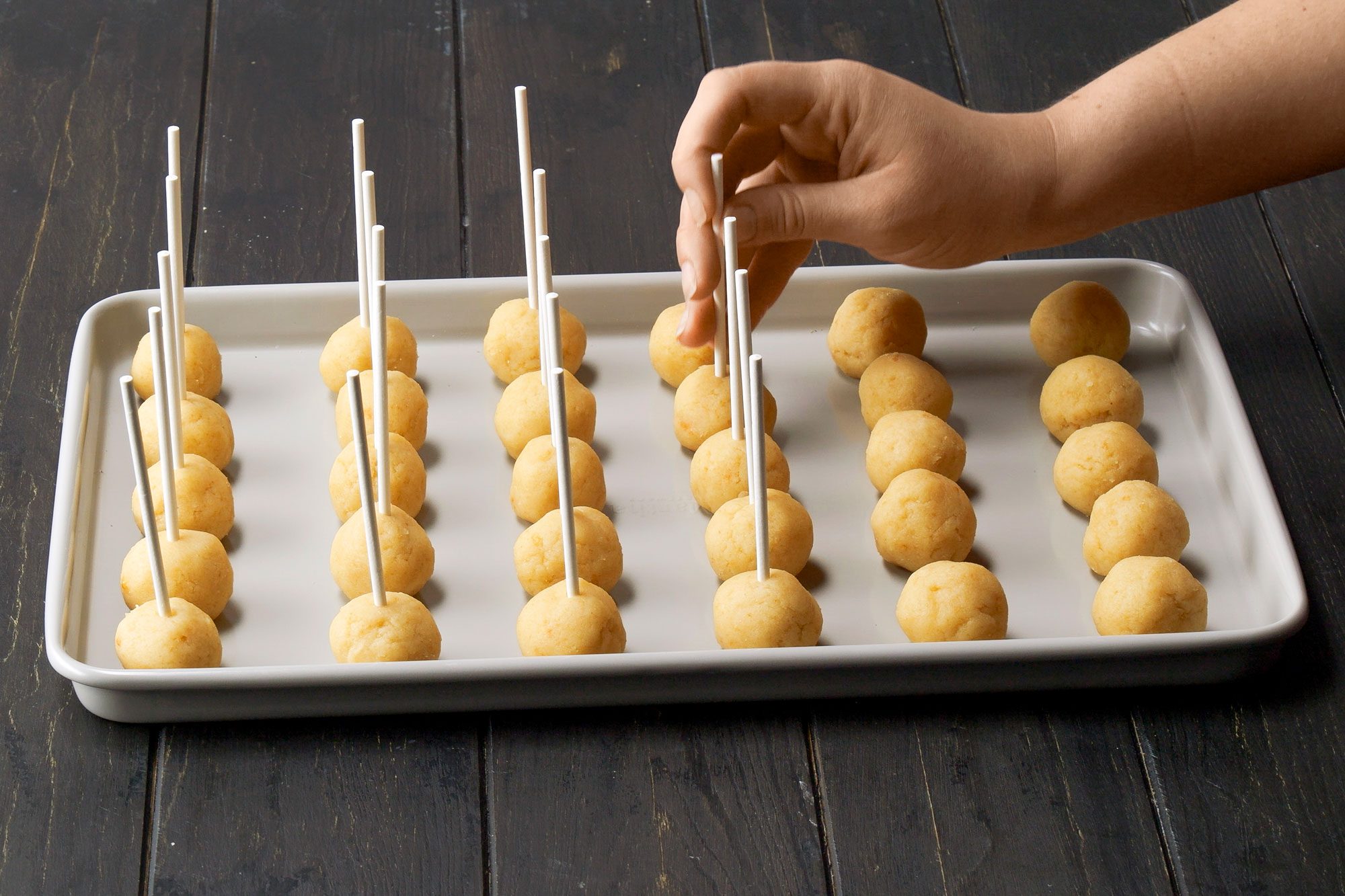 3/4th shot of a white tray filled with round dough balls arranged in neat rows; each ball has a thin white stick inserted into it; a hand is seen placing or adjusting one of the sticks while others are already in place; the tray is set against a dark wooden surface