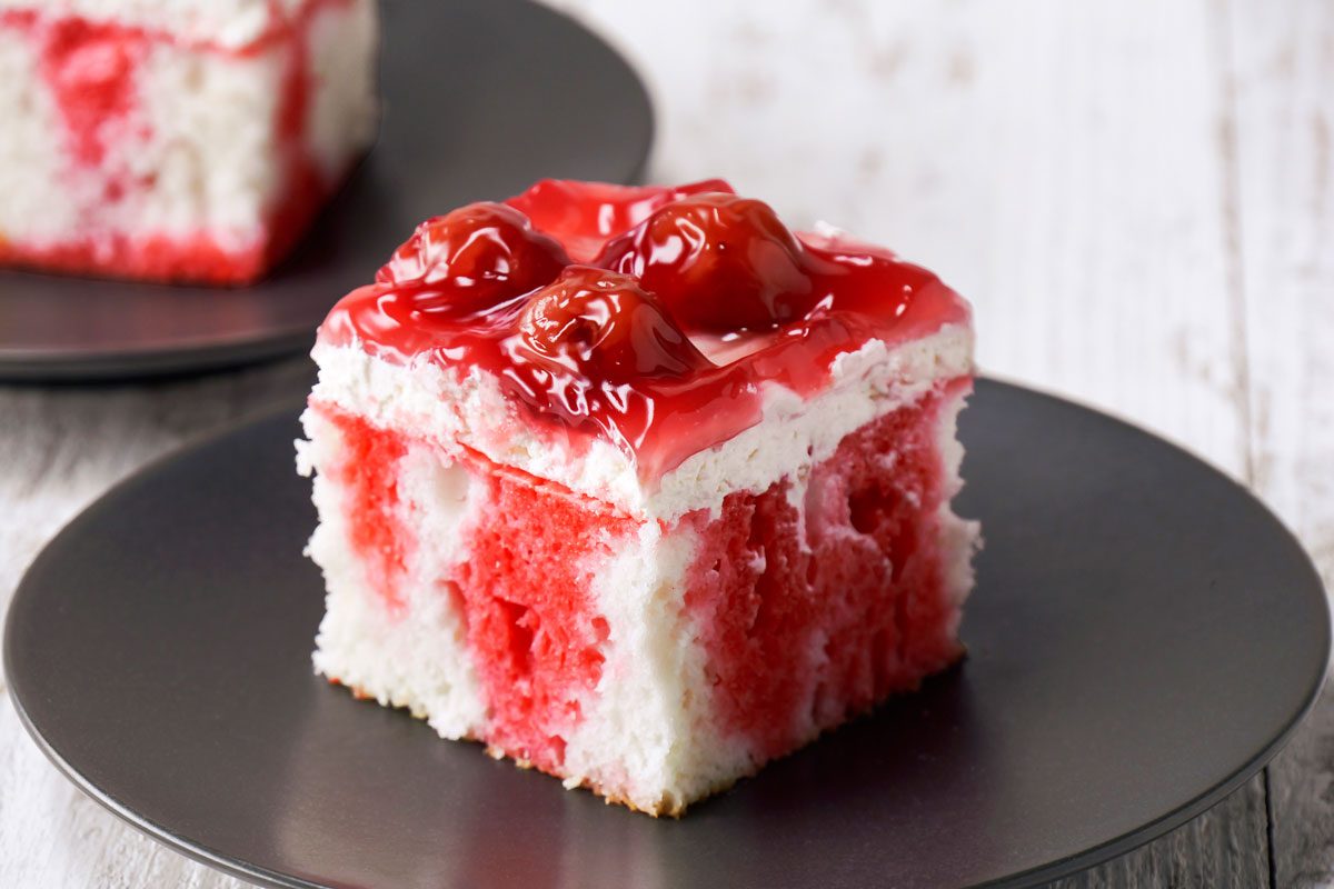 closeup shot of a slice of cherry cake on a black plate; the cake is topped with cherry filling and whipped cream; the cake is sitting on a white wooden surface; there is a second plate in the background with another slice of cake on it