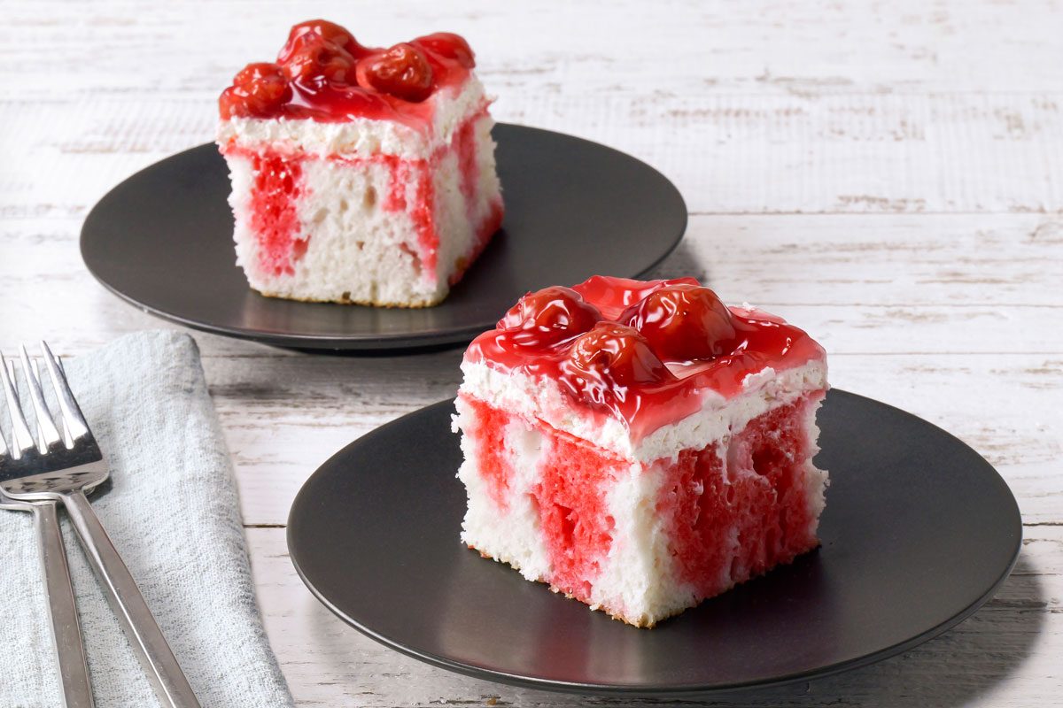 3/4th shot of two pieces of cherry cake on black plates; the cakes are on a white wooden table; the cakes are topped with whipped cream and cherries; the cherries are red and glossy; the cakes are white with pink and red marbling; there is a blue napkin to the left of the cakes, with two forks on top of it