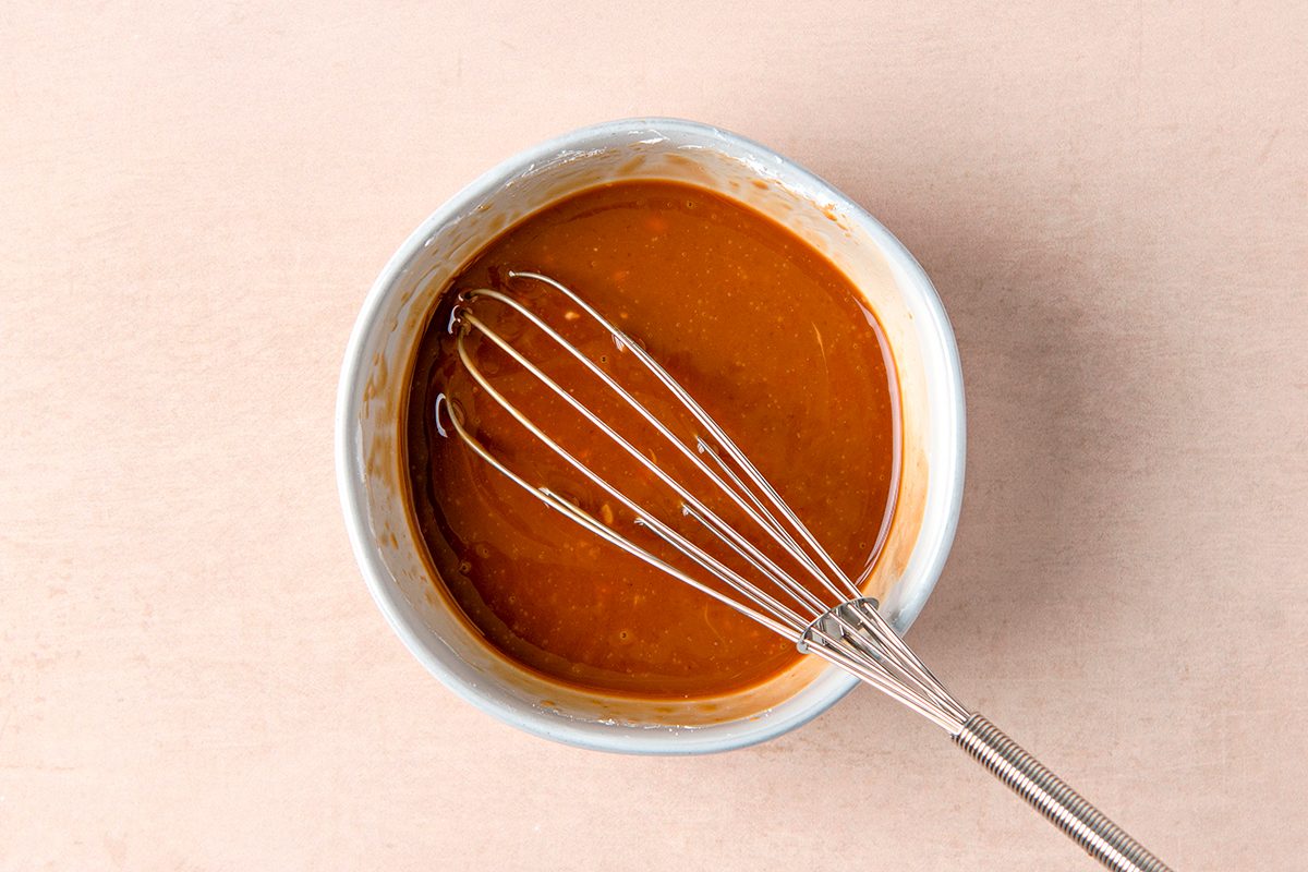 A small bowl filled with caramel sauce sits on a light surface. A metal whisk rests inside the bowl, partially submerged in the smooth, golden-brown caramel. The scene suggests preparation or cooking.