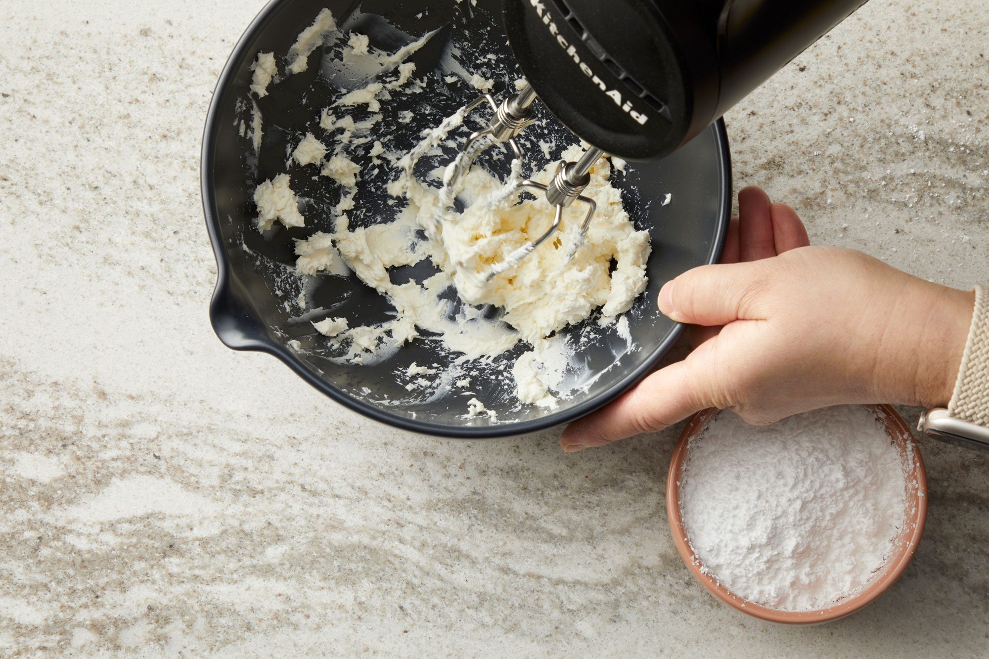overhead shot of a dark gray bowl on a light colored, speckled countertop; inside the bowl, a white, creamy substance, likely cream cheese or frosting, is being mixed with a black KitchenAid hand mixer; partially submerged in the white mixture; a hand is also visible, to the right of the bowl, a smaller, tan bowl contains a white powder, possibly flour or powdered sugar; the countertop appears to be a marble or granite surface